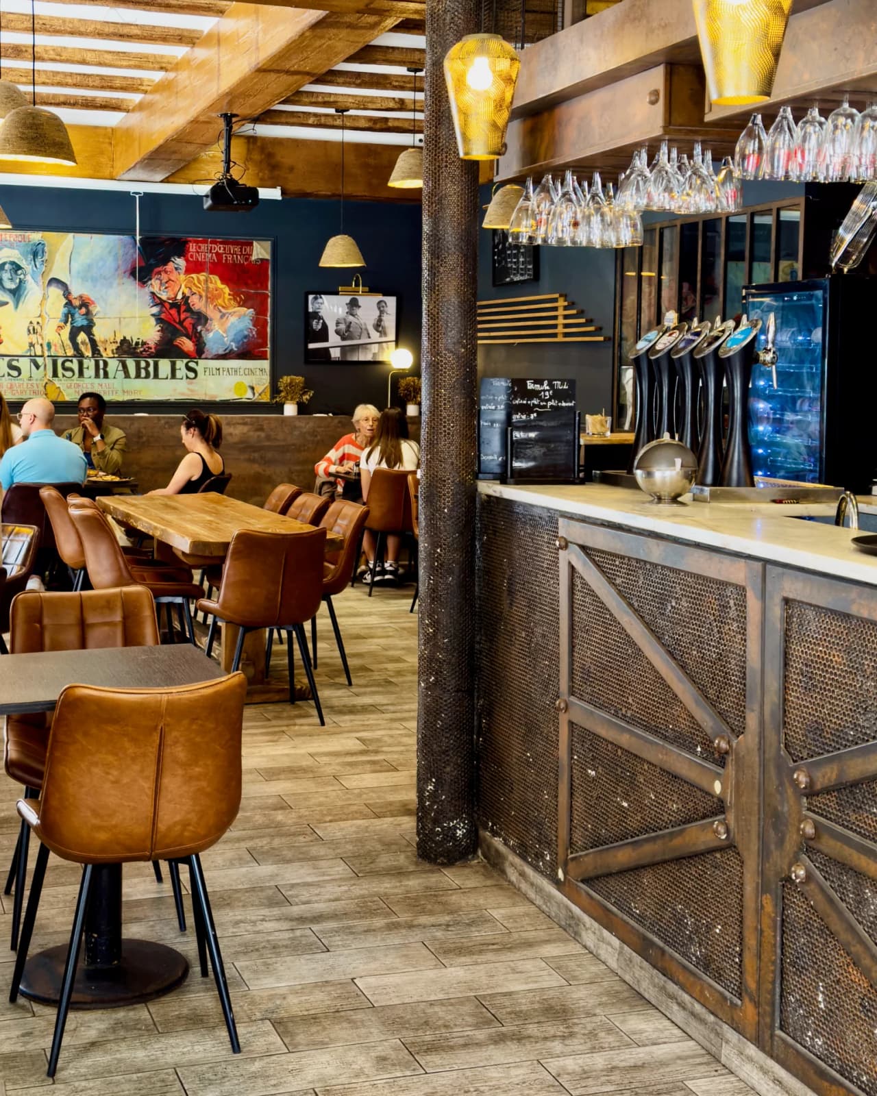 Intérieur moderne et chaleureux de l'Art Bistro à Paris 9ème, montrant une salle avec tables en bois, chaises en cuir camel, un bar avec verres suspendus, poutres apparentes, luminaires dorés et affic