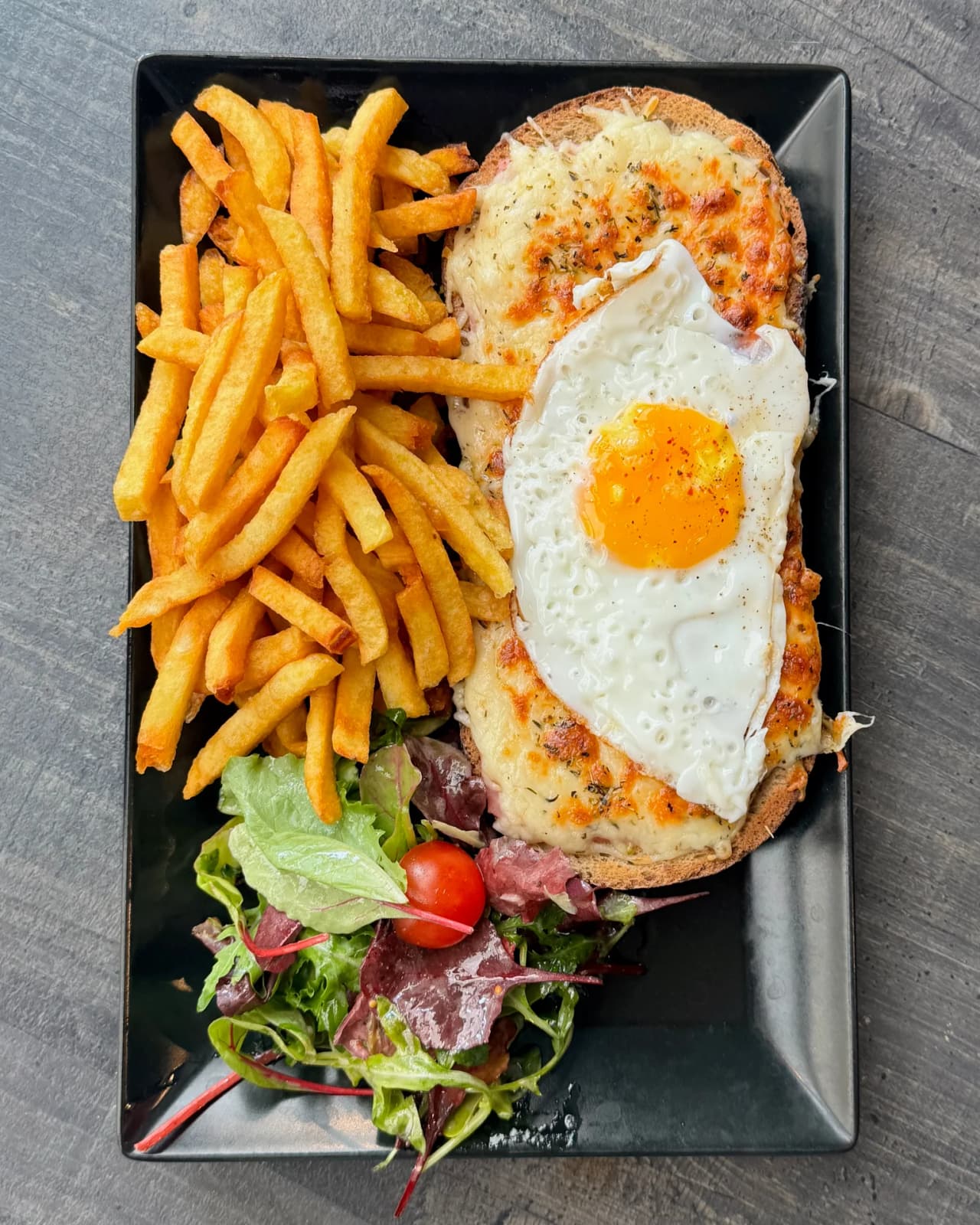 Un croque madame servi sur assiette noire avec un oeuf au plat, accompagné de frites dorées et d'une salade verte avec tomates cerises. Le sandwich est gratiné au fromage fondu.
