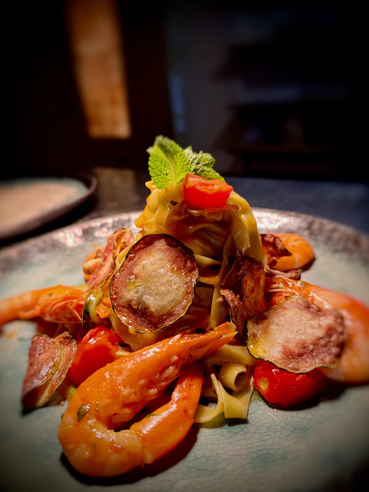 Plat de pâtes fraîches garnies de crevettes roses, courgettes grillées et tomates cerises, décoré d'une feuille de menthe, servi sur une assiette grise texturée