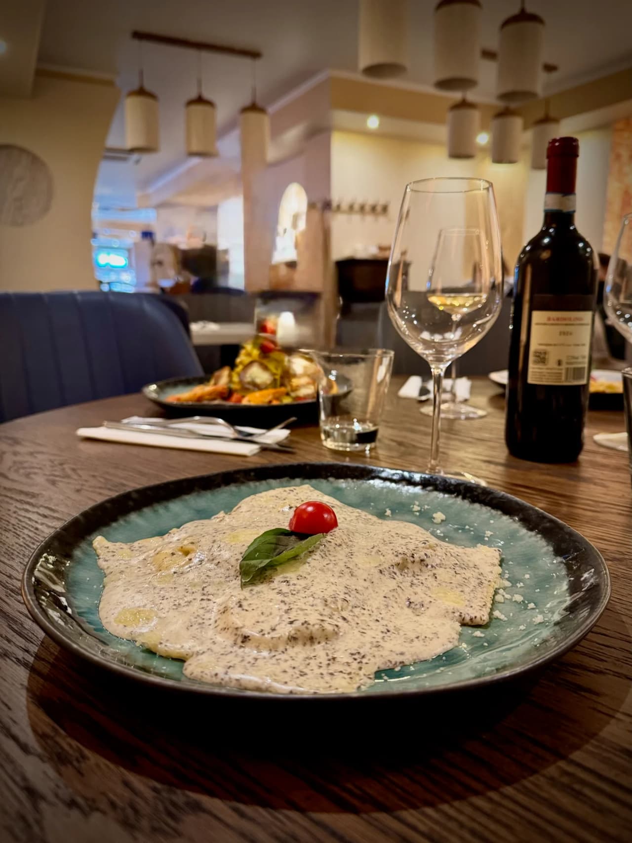 Assiette de raviolis à la sauce crémeuse truffée, décorée d'une tomate cerise et d'une feuille de basilic, servie sur une assiette turquoise. En arrière-plan, une bouteille de vin rouge et des verres