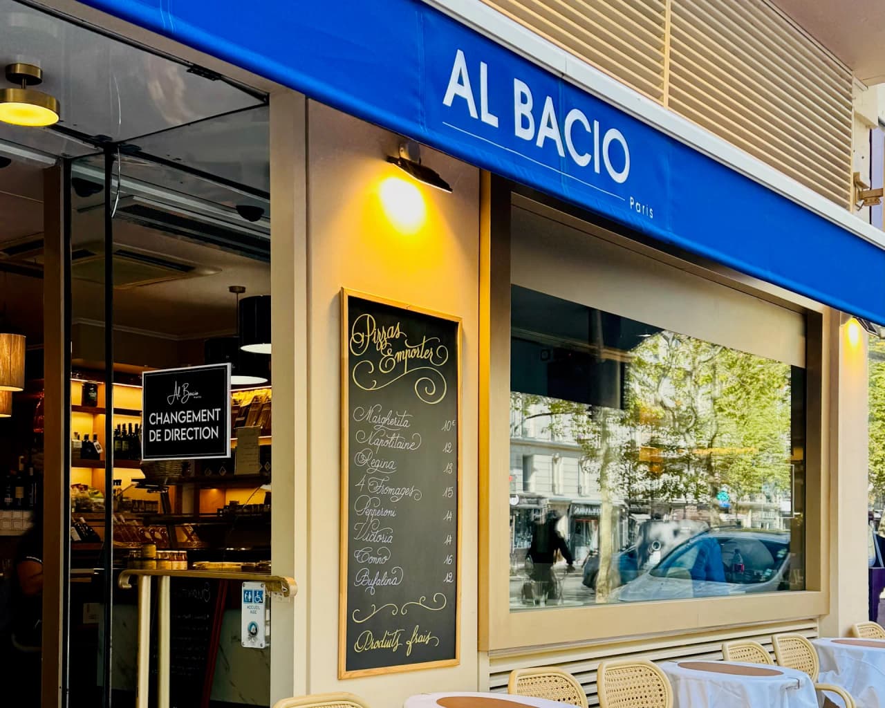 Façade du restaurant Al Bacio avec store bleu, grande vitrine, ardoise de menu manuscrite et chaises en rotin sur la terrasse. Enseigne lumineuse et ambiance chaleureuse avec éclairage jaune.