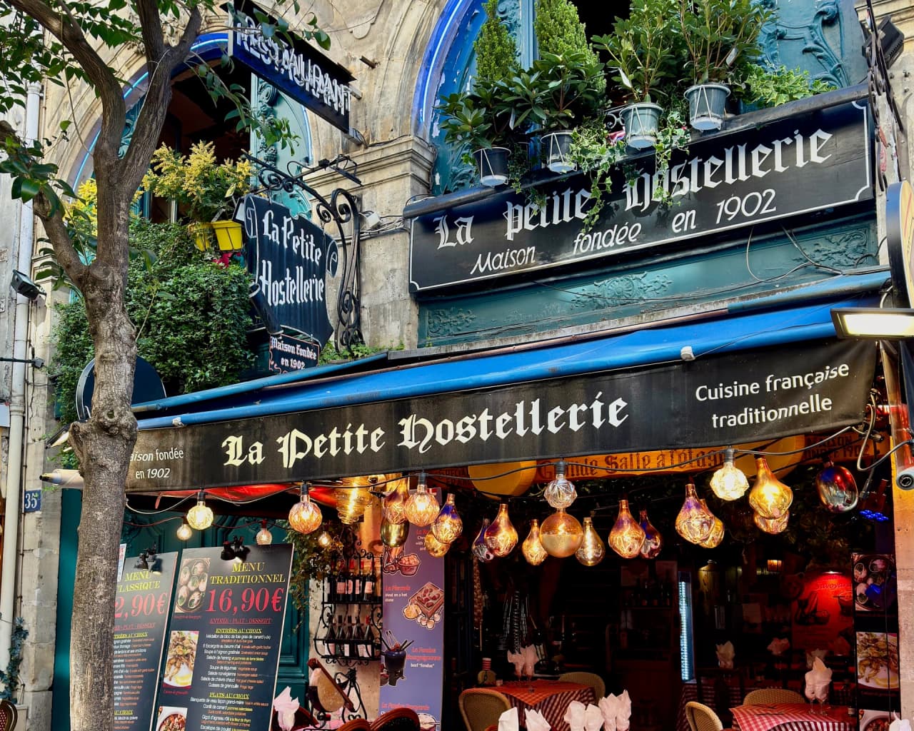 Devanture d'un restaurant traditionnel français avec enseigne gothique 'La Petite Hostellerie' fondée en 1902. Façade ornée de lanternes vintage, végétation luxuriante et menus affichés. Décoration ex