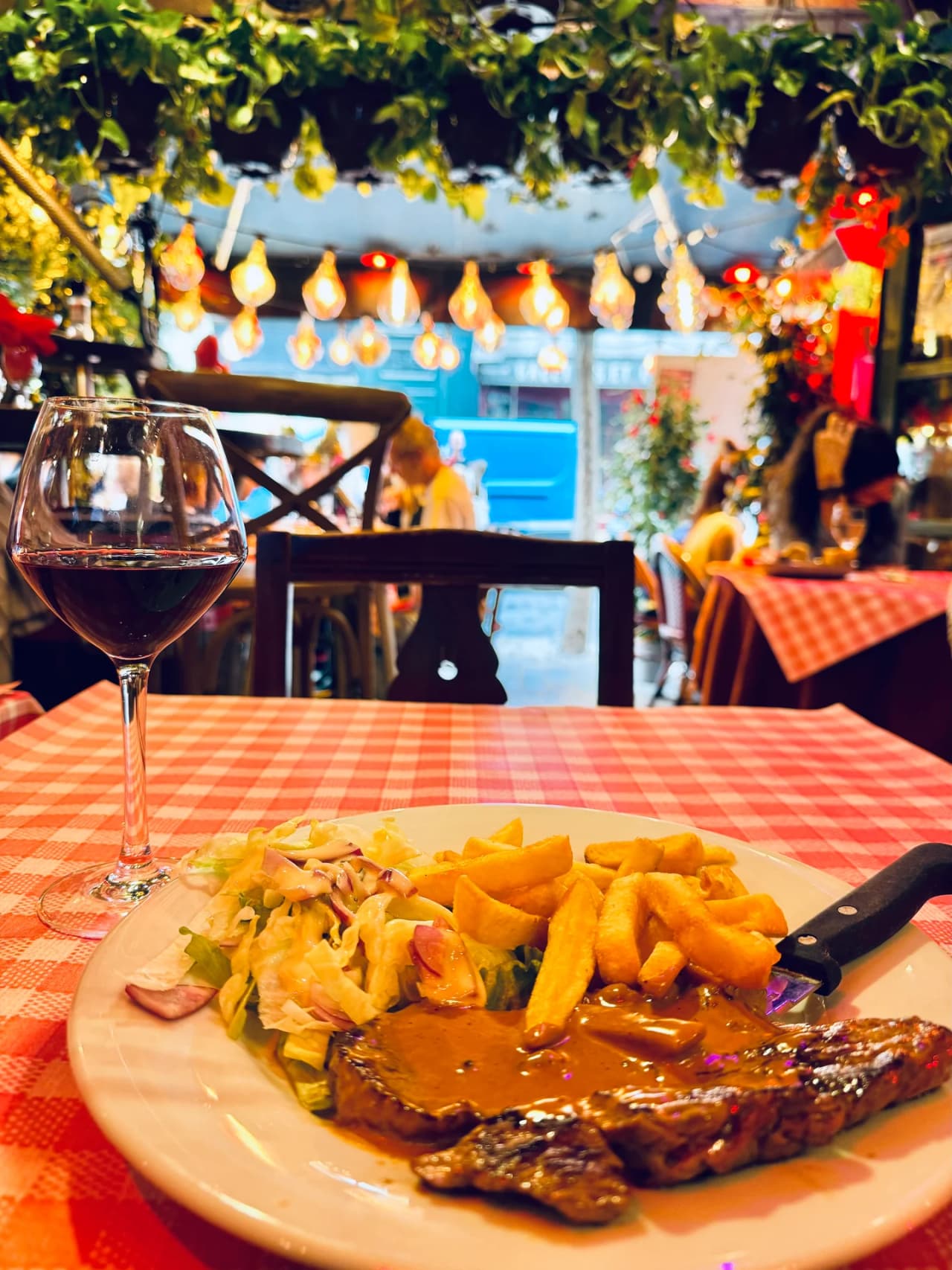 Assiette de steak-frites avec sauce au poivre, salade verte et verre de vin rouge sur une nappe à carreaux rouges et blancs. En arrière-plan, une terrasse couverte décorée de guirlandes d'ampoules et