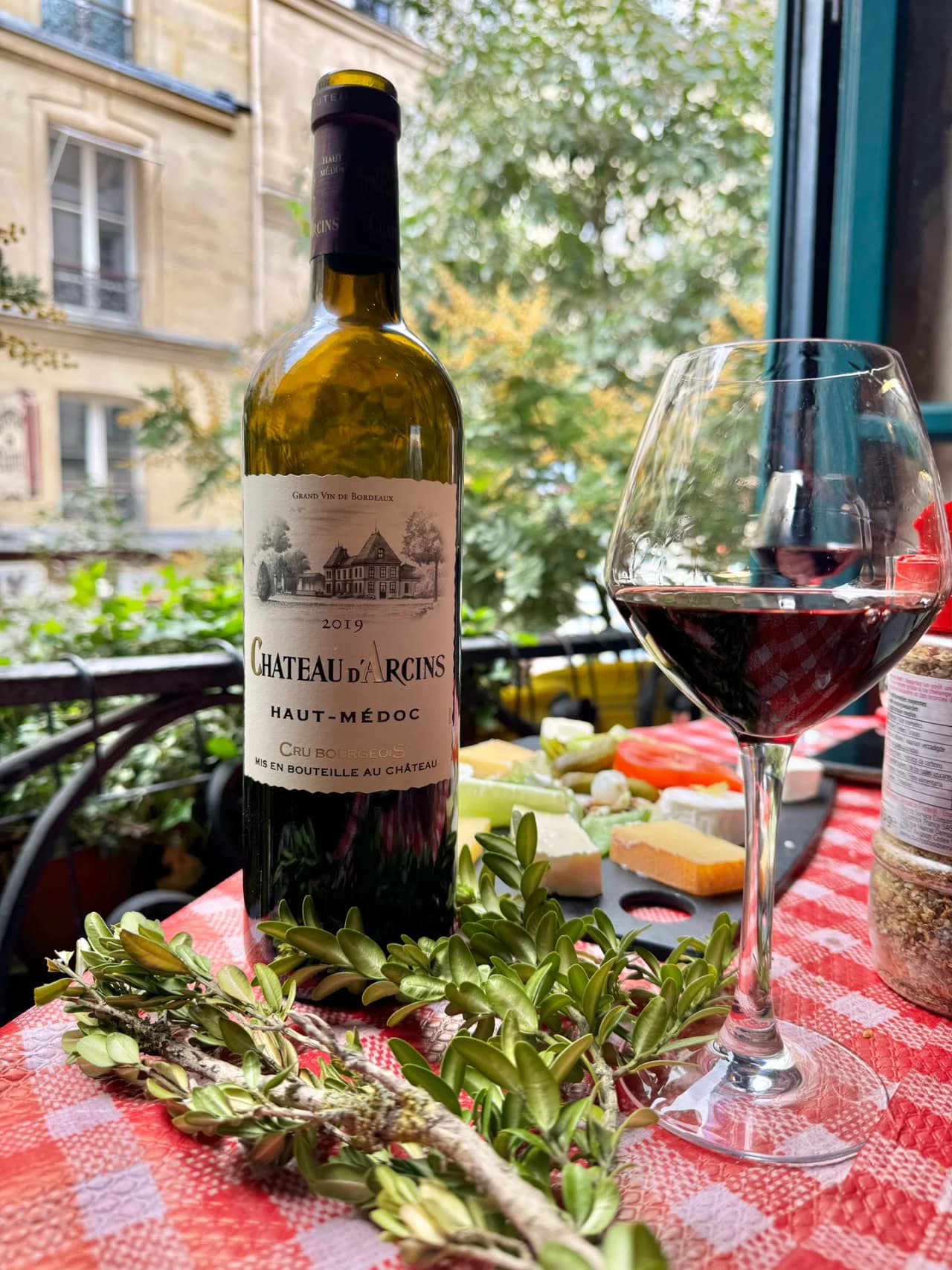 Bouteille de vin Château d'Arcins Haut-Médoc 2019 avec un verre de vin rouge sur une nappe à carreaux rouge et blanc, accompagnée de fromages et décorations végétales, vue sur façade parisienne en arr