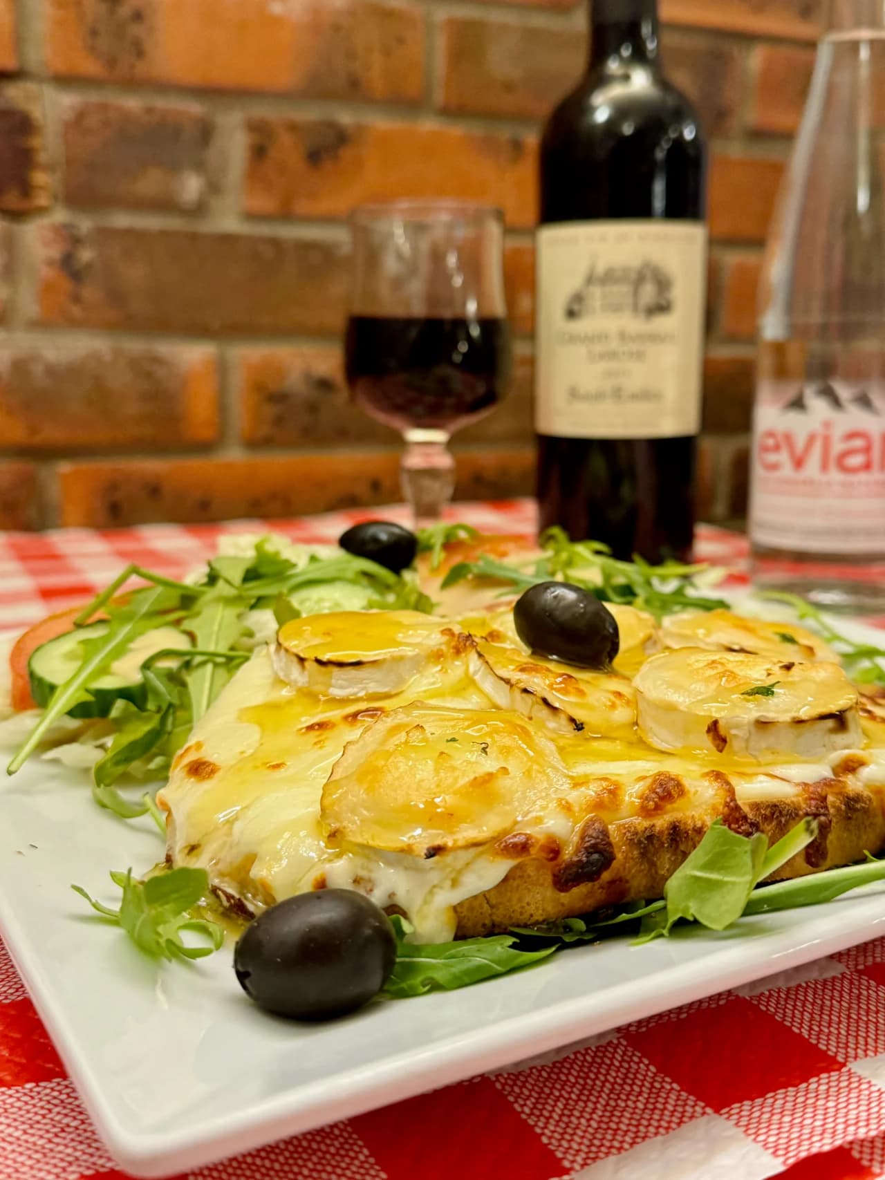 Un croque monsieur gratinée au fromage de chèvre, garni d'olives noires et de roquette, servi sur une assiette blanche carrée. En arrière-plan, un verre de vin rouge et une bouteille sur une nappe à c