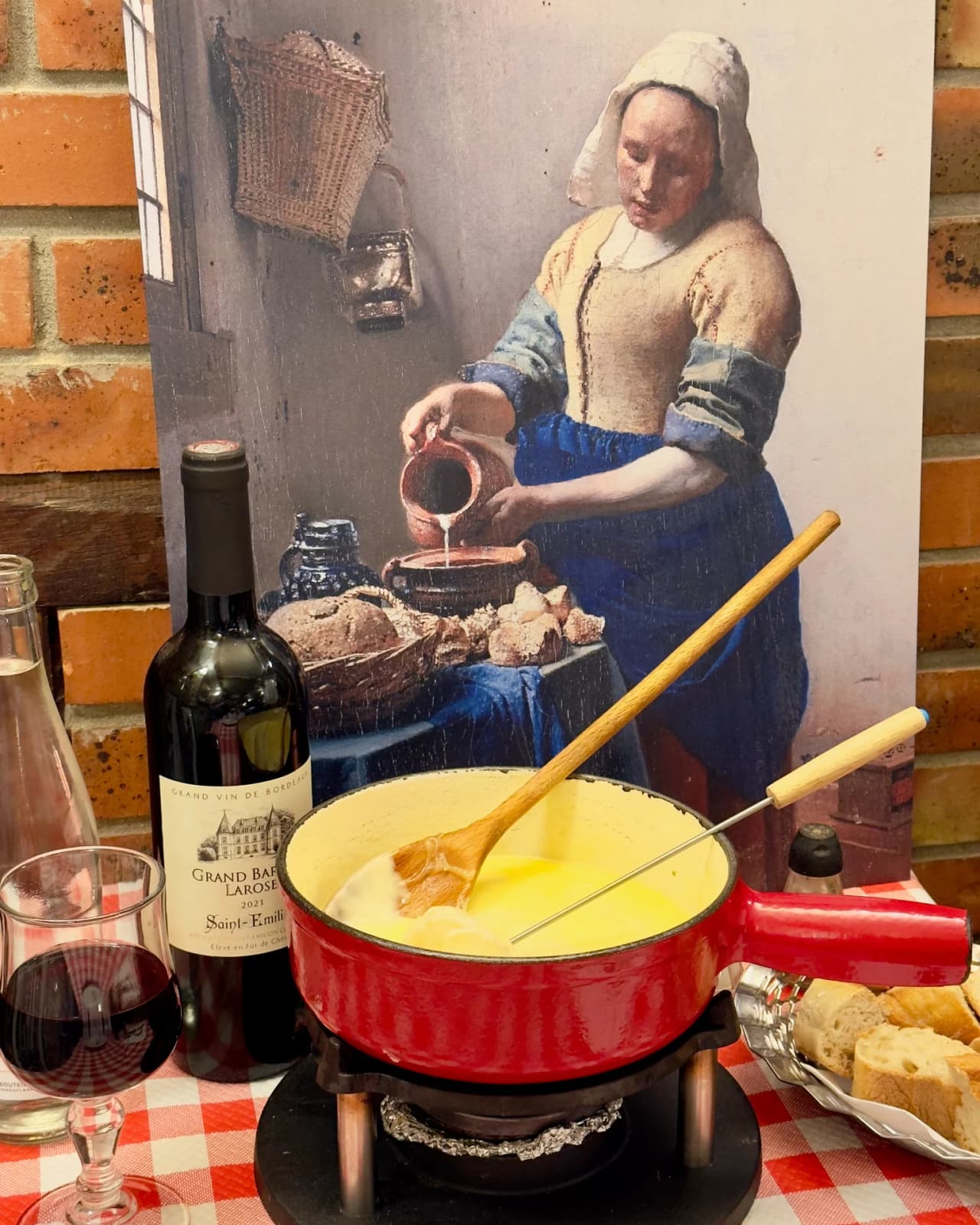 Une fondue savoyarde dans un caquelon rouge sur une table avec nappe à carreaux rouges et blancs. Une bouteille de vin de Saint-Émilion et du pain. En arrière-plan, reproduction du tableau 'La Laitièr