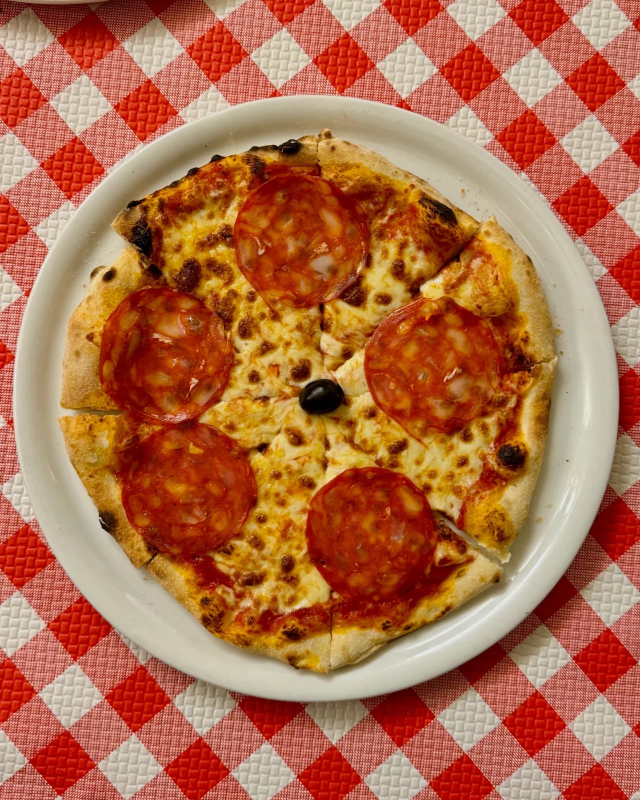 Pizza artisanale garnie de tranches de chorizo et de fromage fondu, servie sur une assiette blanche ronde. Une olive noire décore le centre. La pizza repose sur une nappe à carreaux rouges et blancs t