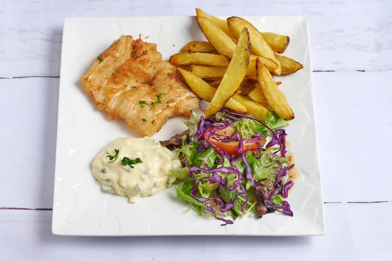 Assiette de fish and chips servie sur fond blanc comprenant un filet de poisson pané doré, des frites épaisses, une sauce tartare crémeuse et une salade fraîche colorée