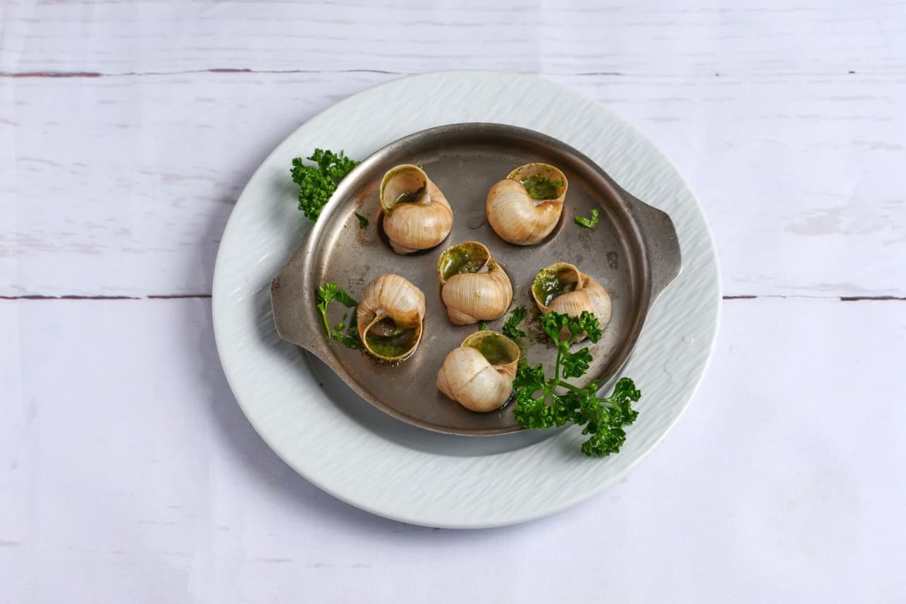 Assiette de six escargots de Bourgogne présentés dans leur coquille avec beurre à l'ail et persil, servis dans un plat traditionnel en métal posé sur une assiette blanche