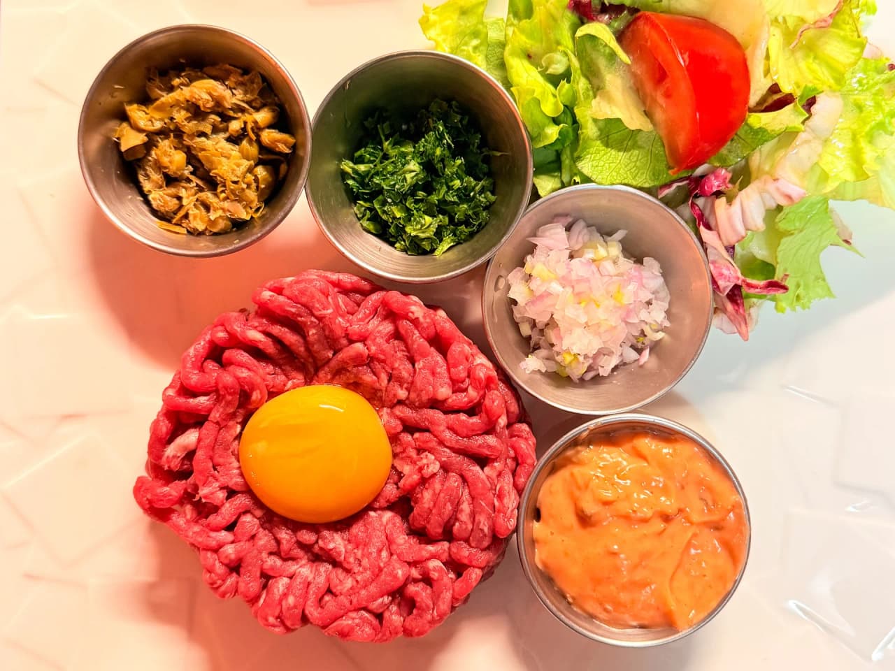 Préparation de tartare de boeuf cru haché finement avec jaune d'œuf au centre, accompagné de petits bols de condiments : échalotes, persil, câpres, sauce et salade verte fraîche
