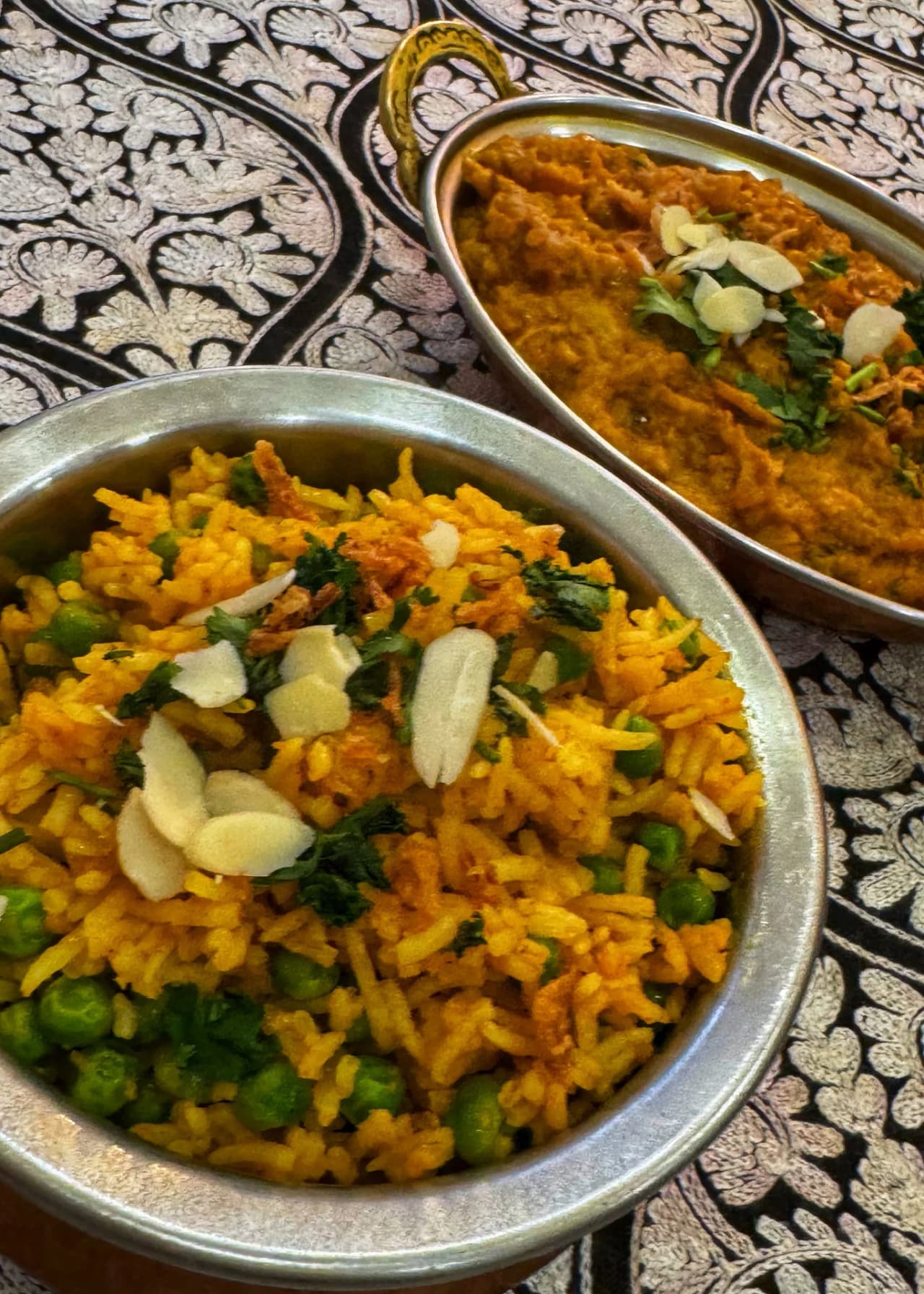 Deux plats indiens servis dans des bols en métal argenté sur un tissu décoratif. Au premier plan, un riz biryani jaune doré garni de petits pois, amandes effilées et coriandre fraîche. À l'arrière, un