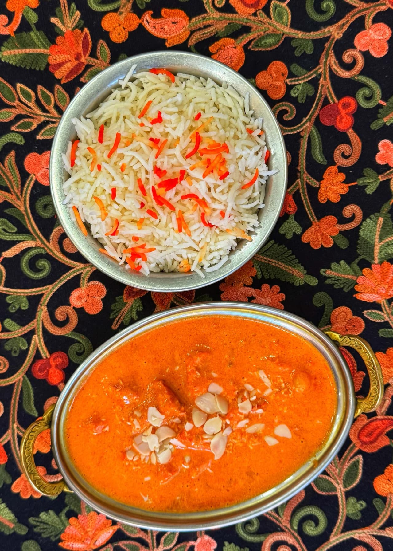 Vue de dessus de deux plats indiens servis sur un tissu brodé traditionnel : un bol de riz basmati blanc parsemé de poivrons rouges et oranges, et un curry orange crémeux garni d'amandes effilées