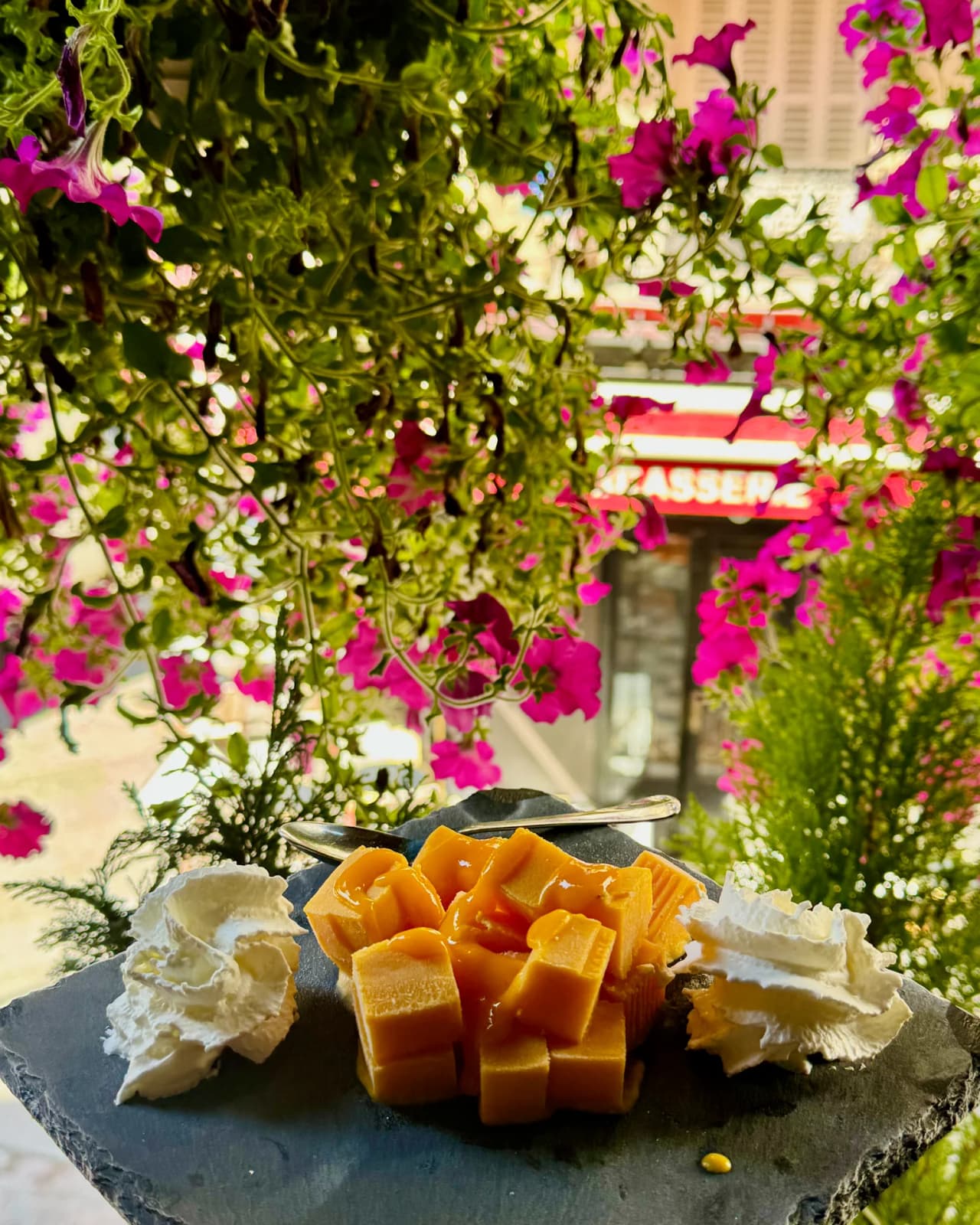 Plateau d'ardoise présentant trois variétés de fromages français sur une table en terrasse fleurie, avec des pétunias roses et blancs en arrière-plan et l'enseigne du restaurant Safran visible