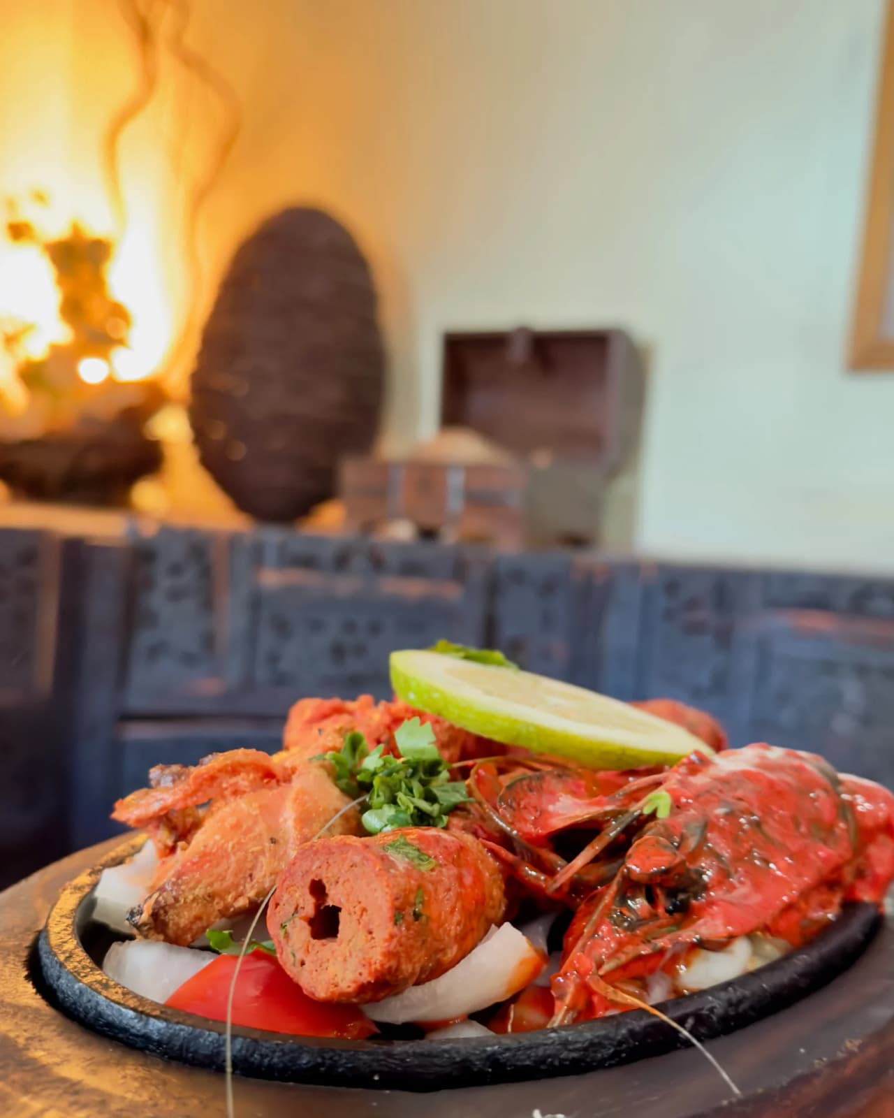 Assiette de fruits de mer avec crevettes frites, calamars et homard sur une planche noire, garnie de citron vert et coriandre fraîche, servie au restaurant Safran avec cheminée en arrière-plan