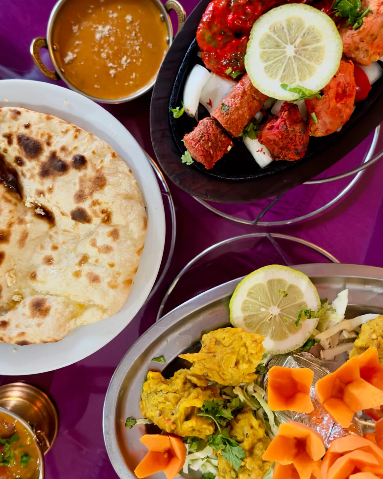 Présentation de plats indiens traditionnels : morceaux de poulet tandoori rouge vif avec citron, pain naan doré, curry safran dans un bol en cuivre, et garnitures colorées de carottes sculptées
