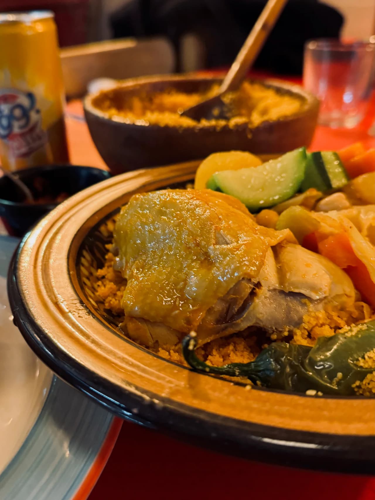 Prato de cuscuz servido num tajine tradicional com frango dourado, acompanhado de legumes frescos (cenouras, curgetes) e sêmola aromática. Um molho é servido numa taça à parte.