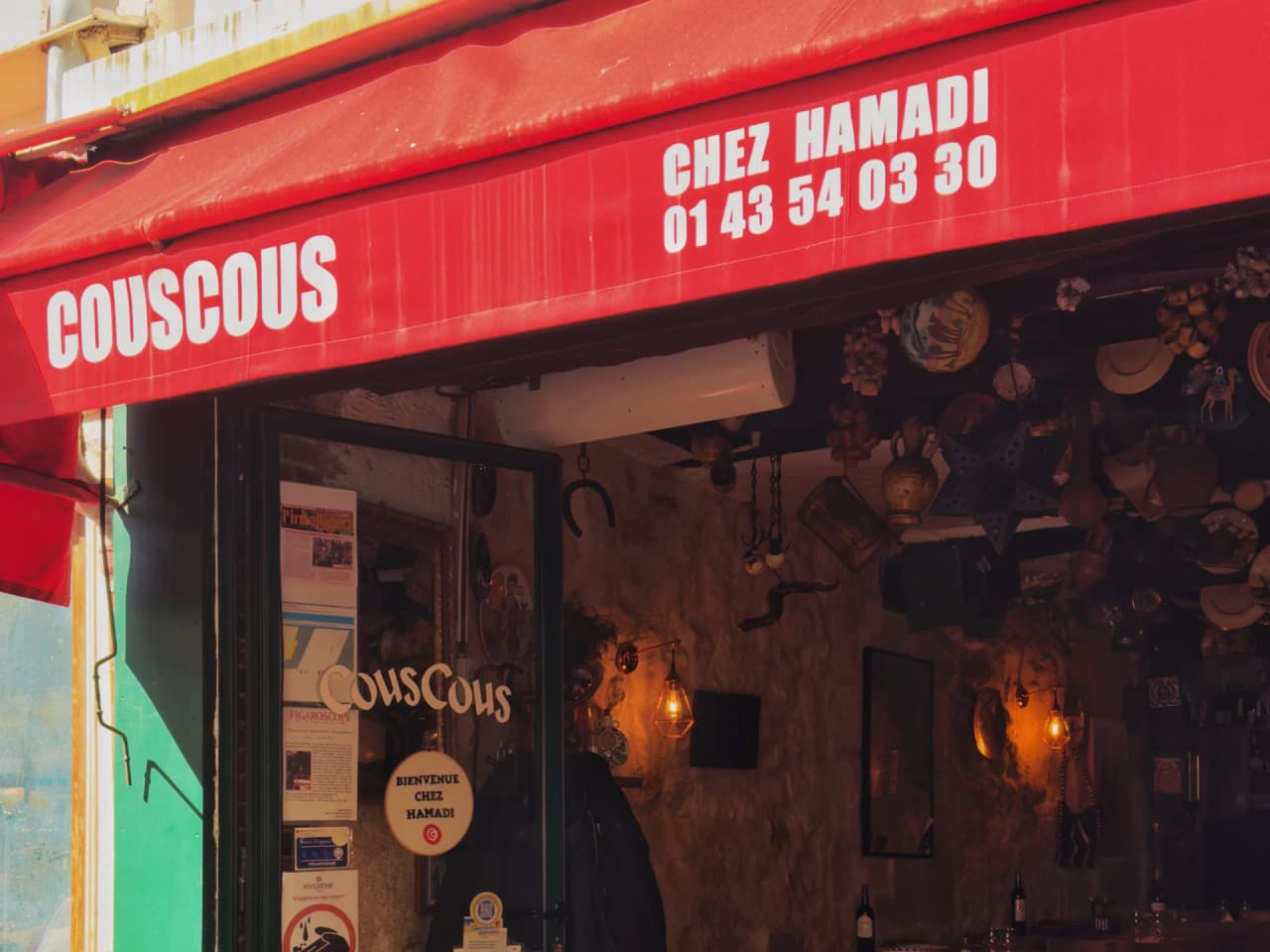 Fachada de um restaurante de cuscuz com toldo vermelho vivo onde se lê 'COUSCOUS' e 'CHEZ HAMADI'. Interior acolhedor visível, com luz suave e decoração norte-africana tradicional nas paredes.