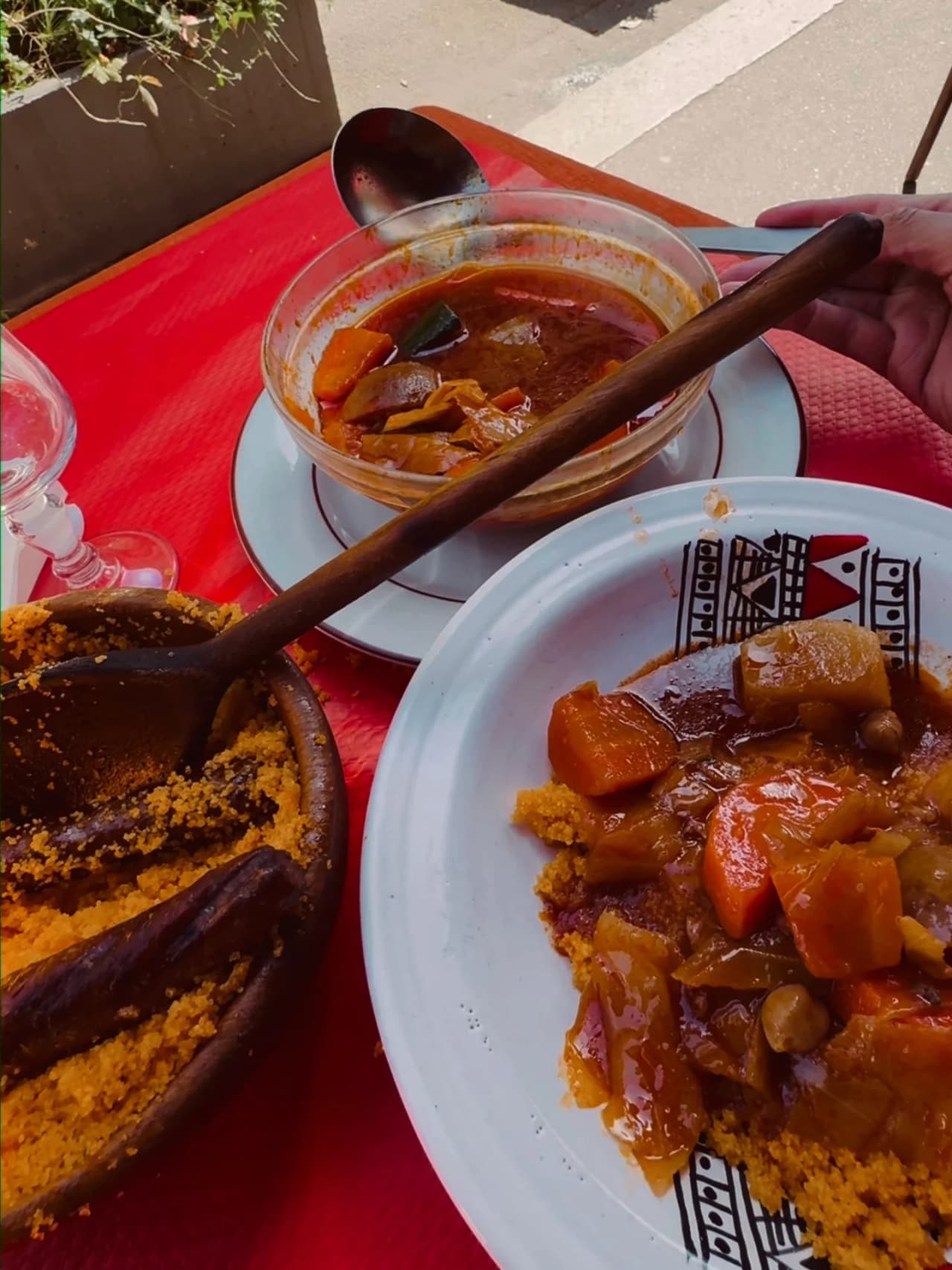 Prato de cuscuz de legumes com molho numa taça de vidro, servido sobre toalha vermelha. Apresenta cenouras, nabos e outros legumes estufados em molho aromático, acompanhado de sêmola.