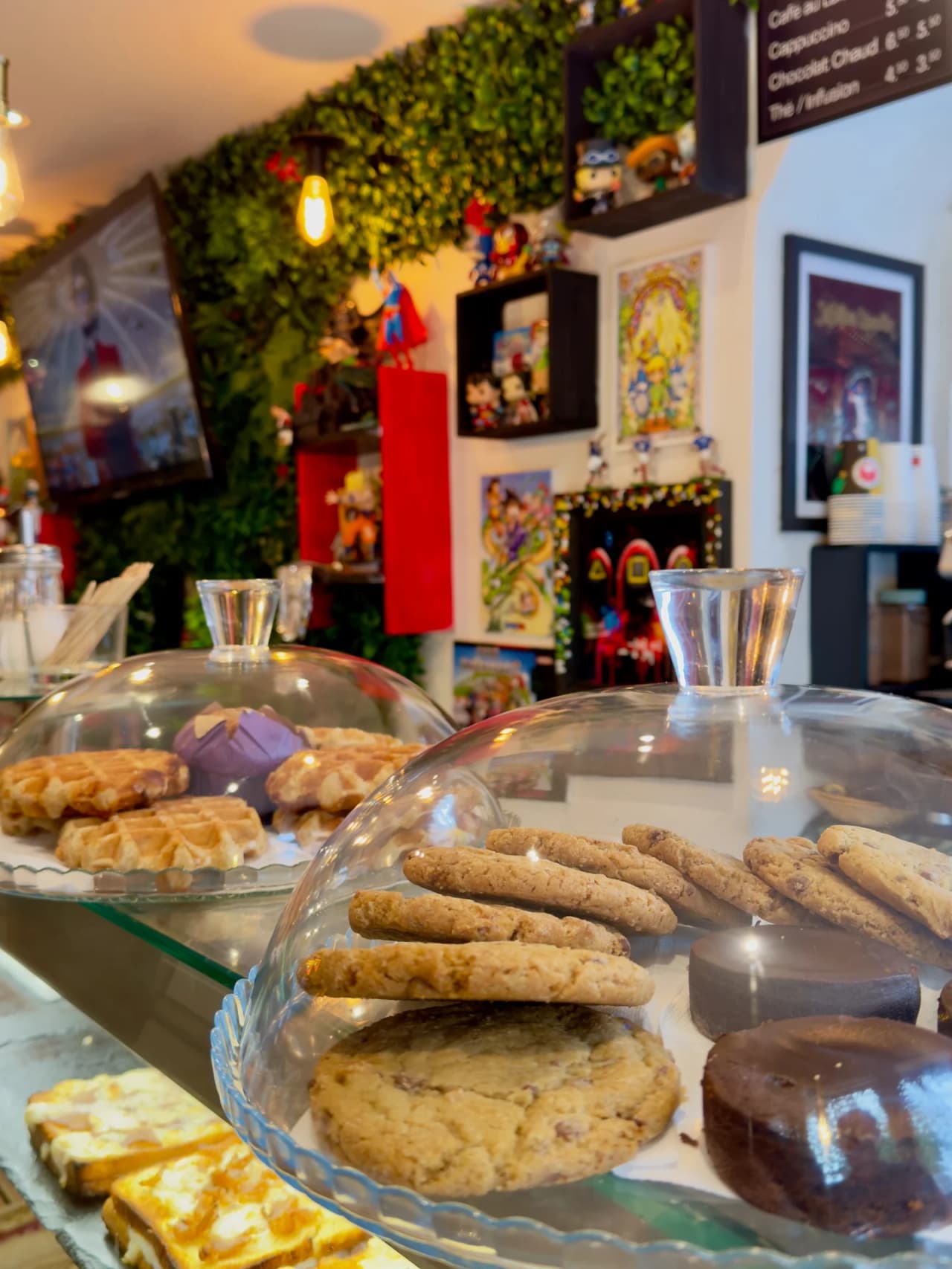 Présentoir de pâtisseries sous cloches en verre montrant des gaufres dorées, cookies et gâteaux, avec en arrière-plan un mur végétal et une décoration thématique manga avec figurines et affiches color