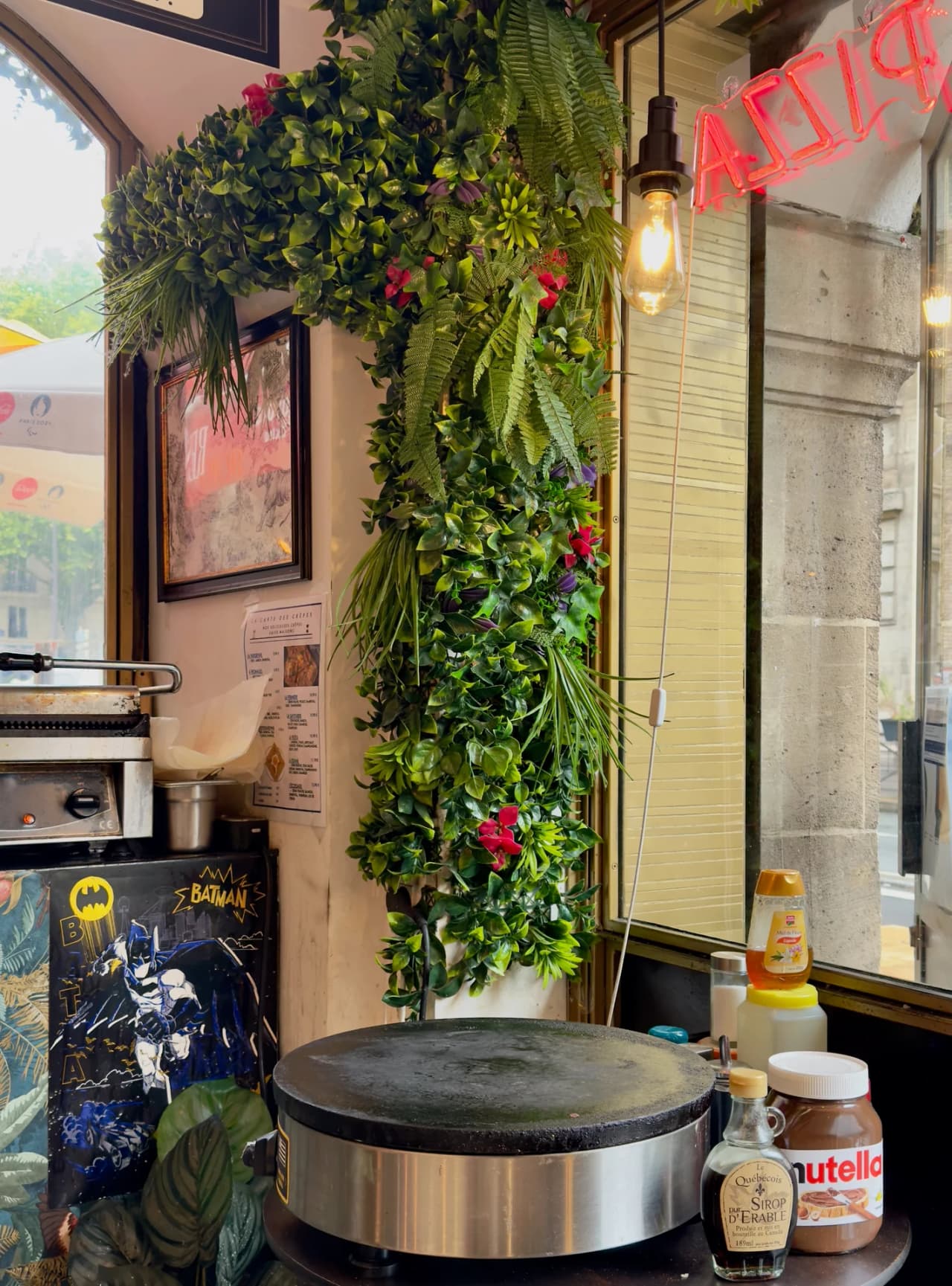 Intérieur d'un restaurant avec une crêpière professionnelle, un mur végétal luxuriant garni de fougères et fleurs rouges, une enseigne néon rouge, et une décoration Batman. Bouteilles de Nutella et si