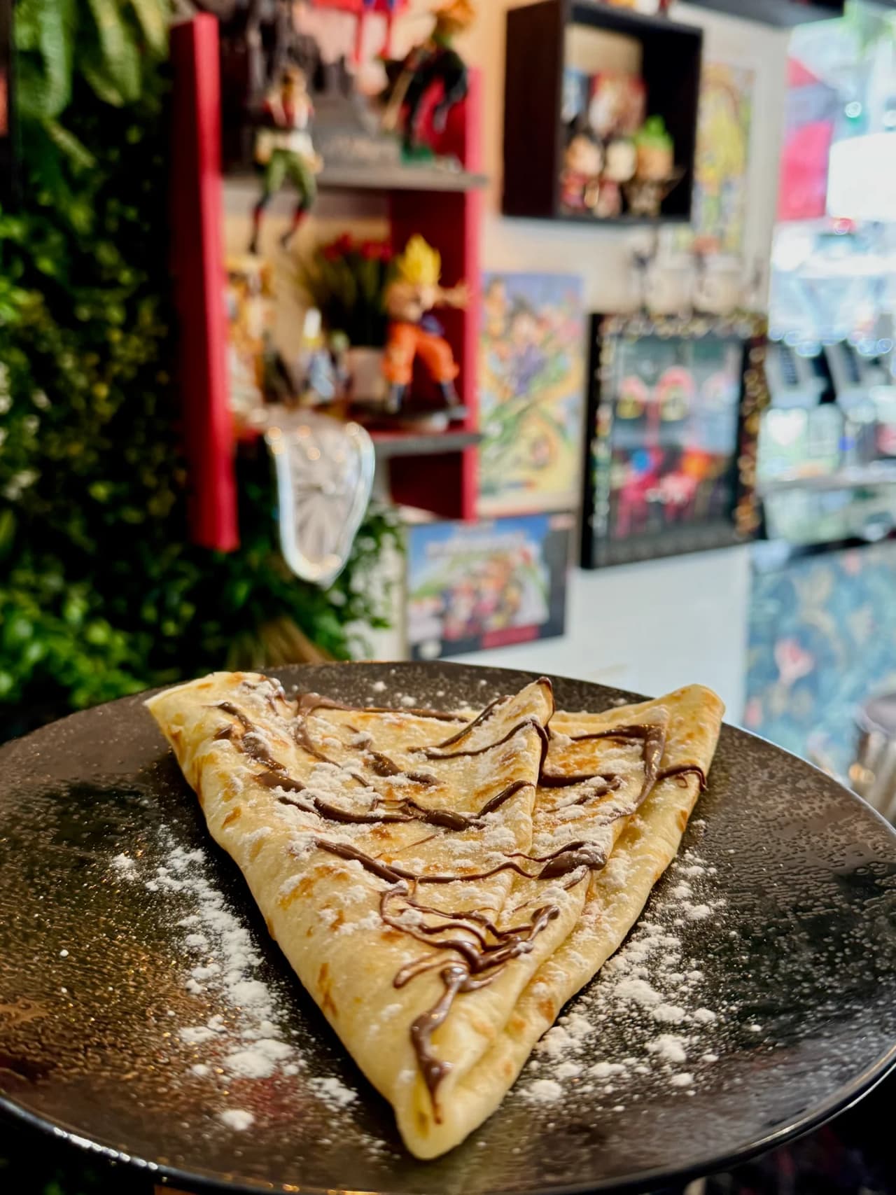 Une crêpe dorée pliée en triangle sur une assiette noire, garnie de filets de Nutella et saupoudrée de sucre glace. En arrière-plan, une décoration colorée avec des étagères rouges et des tableaux mur